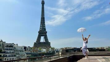 un hermosa joven niña en un verano vestir soportes en contra el fondo de el eiffel torre con un cordón paraguas, ella elevado su mano y como Si tomando apagado allí es un sitio para publicidad viaje agencia foto