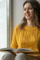 joven hermosa mujer cerca ventana amarillo de punto suéter leer libro foto