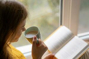 joven hermosa mujer cerca ventana amarillo de punto suéter leer libro foto