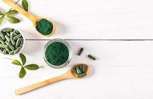 Green powder and spirulina algae tablets in glass jars and spoon on white wooden background. Top view. A copy space. high content of multivitamins. photo