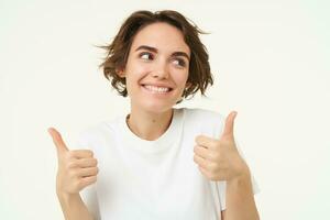 Close up shot of cheerful smiling girl, shows thumbs up, gives approval, likes something, positive feedback concept, stands over white background photo