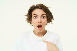 Close up portrait of girl with surprised face, points at herself, looks shocked, stands over white background photo