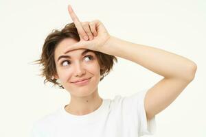 Close up of woman shows loser gesture, l letter on forehead and smiling, mocking, makes fun of someone, standing over white background photo