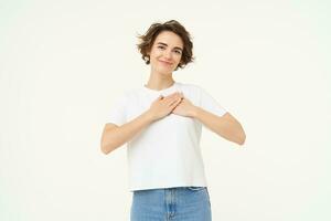 Portrait of brunette woman with hands on chest, looks with care and love, express gratitude, self-care and heart-warming feelings, stands over white background photo