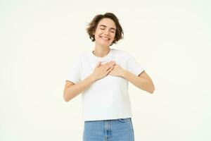 Image of brunette woman holding hands on heart, express gratitude, heart-warming feelings, emotion of happiness, love and care, isolated over white background photo