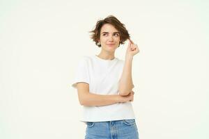 Portrait of thoughtful brunette girl, playing with her curl and thinking with smiling face, standing over white background photo