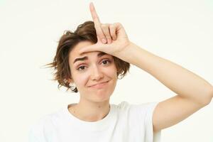 Close up of woman shows loser gesture, l letter on forehead and smiling, mocking, makes fun of someone, standing over white background photo