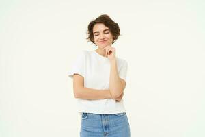 Image of pleased smiling brunette girl, smirks, looks down with confident, satisfied face expression, stands over white background photo