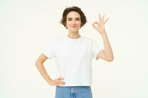 No problem. Portrait of young woman standing in confident pose, shows okay, ok, zero gesture, stands over white studio background photo