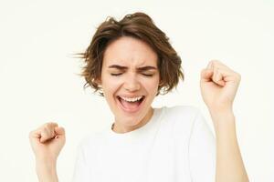 Image of happy, excited brunette woman, clenches her fists, dancing in excitement, feeling amused, standing over white background photo