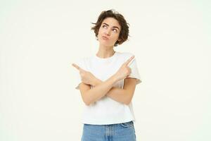 Portrait of brunette woman with thoughtful face, pointing sideways, making choice, decision, standing over white studio background photo