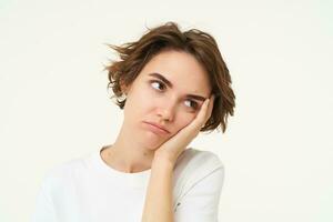 Portrait of young bored woman, leans head on hand and frowns, looks upset and grumpy, thinking, standing over white background photo