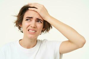 Portrait of young upset woman, slaps her forehead, facepalm, touches head and looks like she is in pain, has headache or migraine, isolated over white background photo