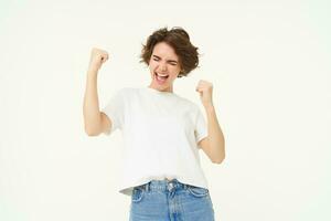 Excited brunette girl, student winning prize, celebrating victory, triumphing, standing in white t-shirt and jeans over white background photo