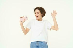 Portrait of carefree girl singing in wireless headphones into smartphone microphone, playing music game on mobile app, standing over white background photo