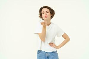 Portrait of young woman sending air kiss, blowing at camera and smiling, standing over white background photo