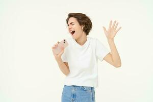 Portrait of happy young woman singing in mobile speakerphone, listening music in wireless headphonse, playing smartphone karaoke game, standing over white background photo