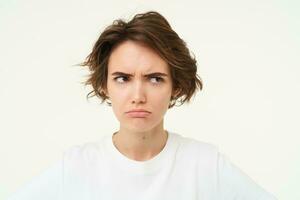 Close up of brunette woman shows angry face, frowning grumpy, pouting and looking disappointed, standing over white background photo