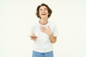 Portrait of young brunette woman texting, sending a message on telephone, using mobile phone and smiling, standing over white studio background photo