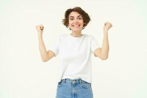 Excited brunette girl, student winning prize, celebrating victory, triumphing, standing in white t-shirt and jeans over white background photo