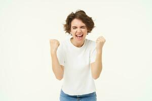 Excited brunette girl, student winning prize, celebrating victory, triumphing, standing in white t-shirt and jeans over white background photo