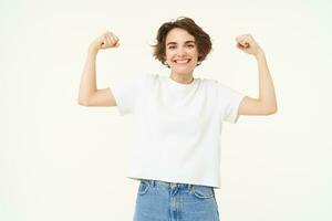 Portrait of happy and confident girl shows muscles, flexing biceps and smiling, posing over white background photo