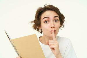 Young woman reads her diary, makes shhh shush face, press finger to lips, hiding something, asking to keep quiet, save secret, standing over white background photo