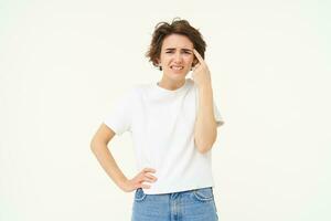 Portrait of woman reacting to smth riddiculous, pointing finger at forehead and frowning, complaining at someone stupid, standing over white background photo