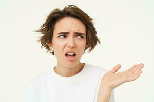 Portrait of disappointed young woman complaining, holding hand sideways, talking, expresses dislike and irritation, standing over white background photo