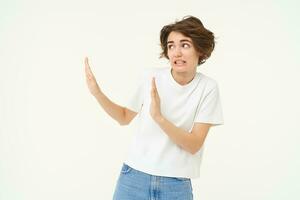 Portrait of awkward, displeased woman shows hand block gesture to the left, avoiding something, refusing, standing over white background photo