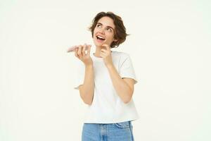 Portrait of brunette woman recording voice message, girl talking on mobile phone, using speakerphone, translating her words, standing over white background photo