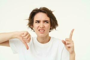 Portrait of disappointed girl, shows thumbs down, pointing at bad product, dislike something, standing over white background photo