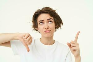 Portrait of sceptical young woman, pointing at something bad, showing thumbs down gesture, posing over white studio background photo