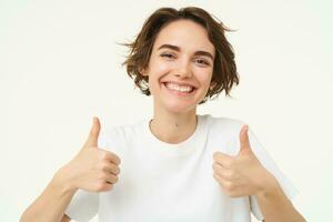 Close up of happy brunette woman, shows thumbs up, approves something, recommends, gives positive feedback, stands over white background photo