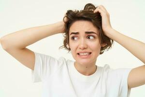 Close up of woman in panic, holding hands on head and clenching teeth, looking troubled, puzzled and frustrated by something, standing over white background photo