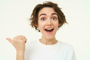 Close up portrait of brunette girl gasps and looks surprised, points left to inquire about promotion, stands over white background photo