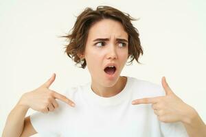 Insulted woman points at herself, being offended or hurt, looks shocked aside, stands over white studio background photo