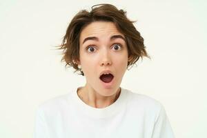 Portrait of woman in shock, drops jaw and stares at camera with mouth wide open, amazed by something, genuine surprise, stands over white background photo