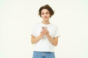 Portrait of brunette girl with smartphone, sending a message, using mobile phone application, smiling and looking happy, white studio background photo