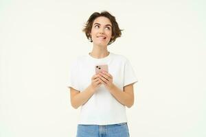 Portrait of brunette girl with smartphone, sending a message, using mobile phone application, smiling and looking happy, white studio background photo