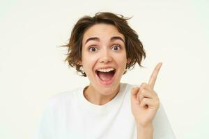 Close up of excited smiling woman, pointing finger up, showing number one, has an idea, thinking of a plan or solution, standing over white background photo
