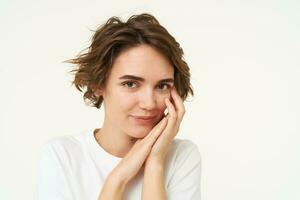 Close up of tender, feminine young woman, touches her face, looks gentle at camera, stands over white background photo