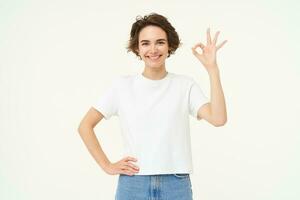 Portrait of woman smiling, showing okay sign with confidence, gives approval, recommends smth good, stands over white background photo