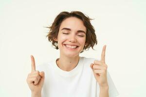 Close up portrait of happy, smiling brunette girl, pointing fingers up, laughing, showing advertisement, standing over white studio background photo