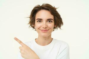 Close up shot of happy, candid woman, laughing and smiling, pointing finger left, showing advertisement, standing over white background photo