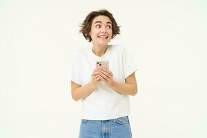 retrato de morena niña con teléfono inteligente, enviando un mensaje, utilizando móvil teléfono solicitud, sonriente y mirando feliz, blanco estudio antecedentes foto