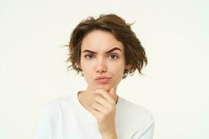 Portrait of thinking woman, touches her chin, looks serious and thoughtful, poses over white background photo