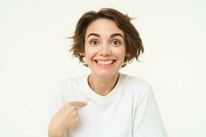 Portrait of girl with surprised face, points at herself amazed, stands isolated over white studio background photo