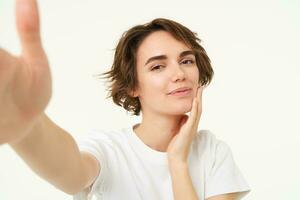 Lifestyle and people. Young woman smiling and taking selfie, posing for photo, holding camera with one hand, standing over white background photo