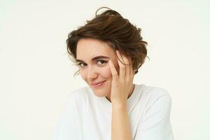 Close up of brunette woman looks shy, covers face with hand and smiles, gazing flirty at camera, stands over white background photo